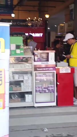 A store entrance is visible with various products displayed outside. A banner with the word "DAN" in yellow is on the left. Next to it is a yellow sign with Indonesian text and a QR code. Inside the store, a person in a yellow shirt and white hat is looking at their phone. There are display cases with various items and signs for brands like Philips. Further into the store, more products are visible. The store appears to sell building materials and home improvement supplies, with signs for TOTO, INFINITI, ASIA TILE, and ANGEL DOORS WINDOWS. Several motorcycles are parked in front of the store. The environment appears to be outdoors with natural lighting.