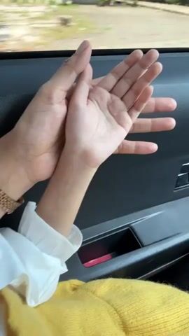 A close-up video shows two hands, one adult and one child, clapping together. The adult's hand is larger and appears to be holding the child's hand. Both hands are positioned in front of a car door and window, with the outside scenery blurred due to motion. The child's hand is wearing a white shirt with ruffled cuffs. The adult's hand has a watch on their wrist. The background shows a blurry view of houses and greenery passing by, suggesting the video was taken while traveling in a vehicle.