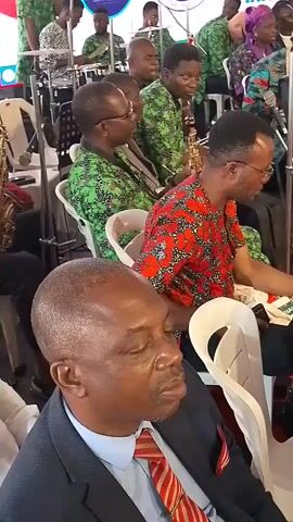 This video shows an indoor gathering in Dushepe, Nigeria, featuring a live band performance. The scene is brightly lit, likely during the day or in a well-lit venue.

**Subjects:** The frame is filled with people, primarily adults. In the foreground, a man in a dark suit jacket, light blue shirt, and red-and-white striped tie is seated, looking down with a contemplative expression. Behind him and to the sides, a band is performing. Many band members are men, wearing vibrant, patterned shirts—some in green and black patterns, others in red and black patterns. Some women are also visible among the performers and seated individuals.

**Activity:** The band is actively playing various instruments. Visible instruments include several saxophones (played by individuals in both green and red patterned shirts) and a trumpet. Drums are also partially visible in the background, along with a drummer. Microphones are set up for the performance. Other people are seated, observing the band.

**Setting and Objects:** The event appears to be a convention or religious gathering, indicated by the large banner in the background. White plastic chairs are used for seating.

**Visible Text:** A prominent banner in the background displays several pieces of text and a logo:
- "11TH ANNUAL CONVENTION IN SESSION"
- A logo featuring a blue map of Africa within a circle.
- Partially visible text below the logo, which appears to say "ENTERING INTO NEW FRUIT GIFTS AND SPIRIT".