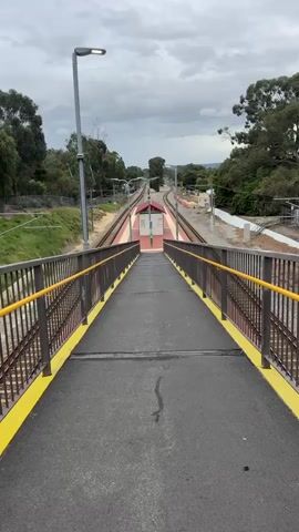 The video shows a view from an overpass looking down at a train station platform. The overpass has a black asphalt surface with yellow painted edges and brown railings. The platform below is painted with red and white stripes and has a small white building with a maroon roof in the center. The tracks extend beyond the platform and curve into the distance. There are streetlights along the tracks and trees surrounding the area. The sky is overcast.