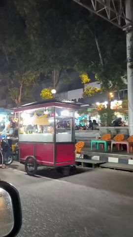 The video shows a nighttime scene of a street lined with food vendors. In the foreground, a person on a scooter is partially visible, facing away from the camera. The scooter is red. To the right, a food cart with a red frame and a white counter is illuminated by lights. The cart has wheels and a sign that says "PADANG". A person wearing a pink shirt is visible behind the counter. Further down the street, another food cart with a blue frame and a white counter is also lit up. Several people are gathered around these carts. In the background, trees and buildings are visible, and streetlights cast a warm glow. The overall atmosphere appears to be that of a bustling night market.