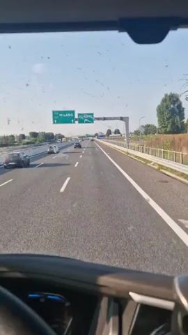 The video is filmed from inside a vehicle looking out through the windshield. The windshield is dirty with smudges and specks. The vehicle is on a multi-lane highway, traveling forward. There are overhead signs indicating directions to "MILANO" and "TURATE." Several cars and at least one white van are visible on the highway. The highway has white lane markings. On the right side of the highway, there is a fence and some vegetation. The sky is clear and blue.