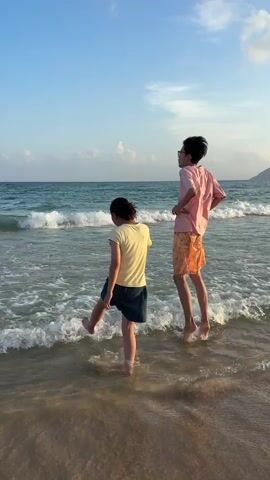 Two people stand in shallow water at a beach. A person with dark hair wearing a yellow shirt and blue shorts faces away from the camera, looking out at the ocean. Another person, wearing glasses, a pink t-shirt, and patterned orange and white shorts, stands to the right of the first person, also facing the ocean. Waves are breaking around their legs. The sky is blue with some clouds.