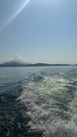 The video captures serene views from a moving boat on a large lake, likely in Binda, Italy, as indicated by the context. The time of day is sunny, with a clear blue sky and bright sunshine.

The first shot (0:00-0:01) shows a wide expanse of deep blue water with a white wake trailing behind the boat. Majestic mountains rise in the distance under a bright sky, with a sunray visible. The boat appears to be moving at a moderate speed.

The scene then transitions (0:01-0:02) to a calmer view of the lake, still showing distant mountains and a small boat in the far distance, suggesting the boat has slowed or is cruising gently.

Subsequent shots (0:02-0:05) offer glimpses of the beautiful surroundings from different perspectives within the boat. One shot (0:02-0:03) looks out through an opening, framing the water and distant shoreline. Another (0:03-0:04) shows the view over metal boat railings, with the lake stretching towards a mountainous horizon, where some distant buildings are visible. The final shot (0:04-0:05) provides a clearer view of the distant shore, revealing buildings nestled among the hills and mountains, giving a sense of a picturesque lakeside community. The water remains calm with gentle ripples, and fluffy white clouds dot the clear blue sky.