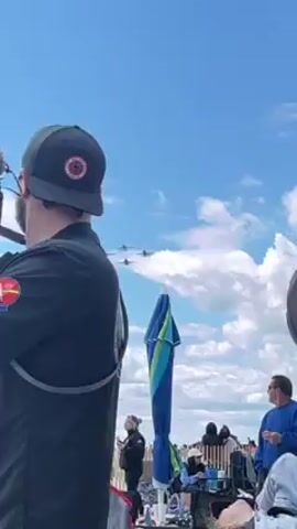 Here is a description of the video:

The video captures four fighter jets soaring through the sky. The planes are silver in color with orange and black accents. They fly in a diamond formation against a backdrop of a blue sky dotted with puffy white clouds. In the foreground, there are glimpses of two individuals, one wearing a black shirt and baseball cap with a patch, and another in a gray hooded shirt. It appears the video was recorded from the ground, with the camera tilting upwards to follow the jets as they pass overhead, resulting in a final shot dominated by the sky and clouds.