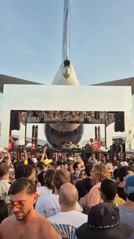 A concert crowd gathers facing a stage with a DJ performing, with a large passenger airplane directly behind the stage. The tail of the airplane, with its characteristic vertical stabilizer, is prominently visible above the stage structure, partially obscuring the view of the clear blue sky. The stage is framed by a white rectangular structure, and there are visible audio speakers and lighting on the stage. The crowd is diverse, with individuals raising their hands in the air. Some people are wearing hats and sunglasses. A man in the foreground on the left has no shirt, and there is a fan in the right foreground. In the distance to the left, another smaller aircraft can be seen.