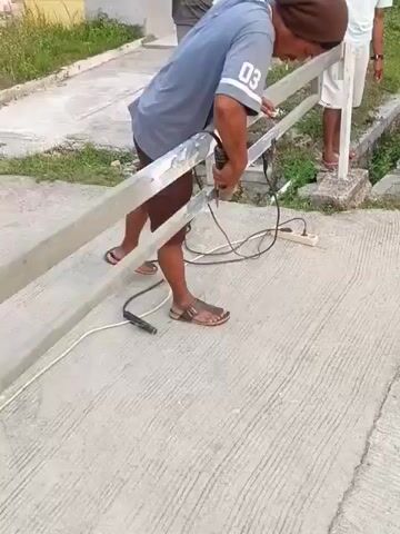 A man in a grey t-shirt and brown shorts is using a handheld grinder on a metal railing. He is standing with his left foot forward and his body bent over the railing. A black power cord runs from an electrical outlet to the grinder. Two other men are standing nearby, watching. One man, in a grey t-shirt, has his arms crossed. The other man, in a white t-shirt, is looking at the railing. The scene appears to be outdoors on a paved surface, with some greenery and houses in the background.