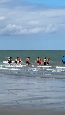 A group of eight people, appearing to be children and young adults, are standing in the shallow water at a beach. The water is calm with small waves gently breaking on the shore. The sky is blue with scattered white clouds. The people are dressed in swimwear and some are interacting with each other in the water. The camera is positioned at a low angle, looking towards the ocean.