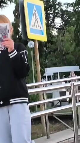 A young girl with blonde hair pulled back in a ponytail is standing outside, holding up a phone to her face. She is wearing a black bomber jacket with white stripes on the collar and cuffs, and light gray pants. The phone screen displays a graphic of a wolf. Behind her, there are green trees and foliage. To her right, a yellow sign with a blue symbol is visible. The girl appears to be looking at the phone. She then lowers the phone and turns slightly to her left, leaning on a metal railing.