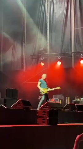 A man is playing a guitar on a stage. He is wearing a dark blue shirt and dark pants. The stage is lit with red, purple, and yellow lights. There are speakers and other equipment on the stage. The man is standing on top of a stack of speakers. There is a large black curtain behind him. Smoke or fog is visible on the stage.