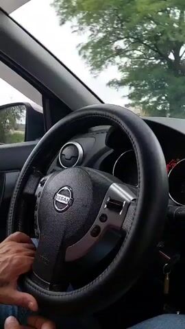 The video shows a first-person perspective from inside a car, focusing on the steering wheel. A person's hands are on the steering wheel, indicating they are driving. The interior of the car, including part of the dashboard and air vents, is visible. Outside the car, through the windshield and side window, trees and a cloudy sky can be seen, suggesting the car is on a road. Audio includes spoken words, which appear to be directions or information related to the drive.