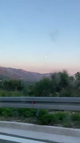 A static shot from a moving vehicle shows a mountain range under a pale blue and pink sky. A full moon is visible in the upper center of the frame. The foreground is blurred, with green foliage and a silver guardrail. In the middle ground, more greenery and some buildings are visible on the slopes of the mountains. The sun appears to be setting, casting a warm glow on the mountain peaks.