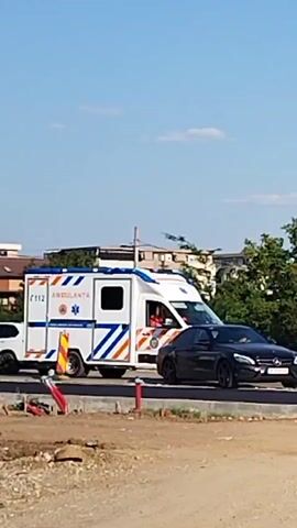 A white ambulance with blue and orange stripes is parked on a road. A black Mercedes-Benz sedan is parked in front of the ambulance. The ambulance has the number 112 and the word AMBULANTA written on its side. There are some traffic cones near the ambulance. The sky is blue with some clouds. The environment appears to be an outdoor area, possibly a construction site or a road under repair, with some trees and buildings in the background.
