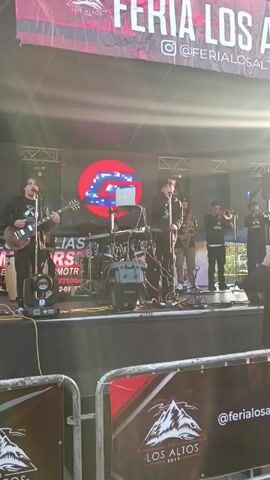A video shows a band performing on a stage during the “Feria Los Altos” event. The band consists of a lead singer with dark curly hair wearing all black and singing into a microphone, a guitarist in a black shirt playing a dark-colored guitar, a drummer behind the drums, a saxophone player, and two trumpet players. The lead singer stands in front of a keyboard and laptop. There is a large screen behind them displaying graphics. The stage is elevated and barricaded with signs with the event logo. The event banner is visible above the stage with “FERIA LOS ALTOS” and the event's Instagram handle. The lighting is bright and sunny.