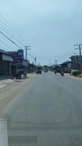 The video presents a first-person perspective from a vehicle moving along a paved road in RawangItiek, Indonesia, during what appears to be a bright, sunny day. The sky is clear, with no clouds visible.

The road is a two-lane street, lined on both sides by various buildings that suggest a mix of residential and commercial establishments. Numerous utility poles with a dense network of overhead electrical wires are prominent along the roadside.

Traffic on the road includes several motorcycles and at least one truck. In the foreground, a person on a motorcycle, wearing a light-colored top and a helmet, is riding away from the camera. Other vehicles are visible further down the road, indicating active daily movement.

On the left side of the road, a blue sign for "BERKAH LAUNDRY" is clearly visible, advertising a laundry service. Below this, another sign is partially obscured. The general scene depicts a typical street in a town or village, bustling with everyday activities.