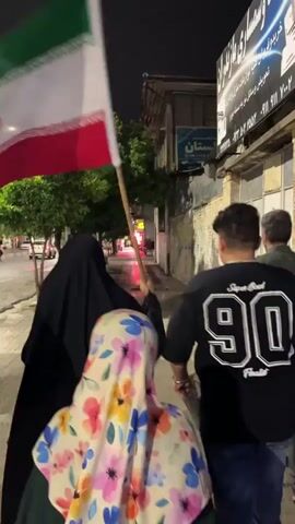 A group of four people walk on a sidewalk at night. The person in the foreground on the right wears a black t-shirt with the number 90 on the back. A person in front of them wears a floral hijab and a green dress. To their left, a person in a black hijab and dress walks while holding an Iranian flag. Another person in a black hijab and dress is visible further to the left, holding a sign. The sidewalk is next to a street with trees and buildings.