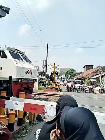 Here's a concise description of the video:

The video shows a train crossing a street-level railroad crossing. The train is carrying tanker cars labeled "PERTAMINA." The crossing is protected by a gate and flashing lights, with people on motorcycles waiting for the train to pass. The day appears to be sunny.
