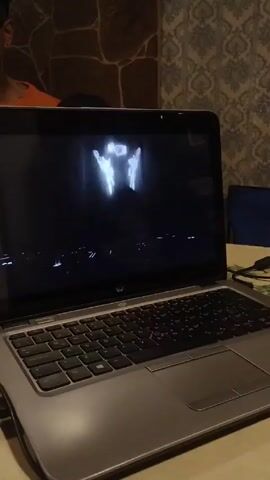 A laptop is on a table displaying a video of a DJ. A person is seated behind the laptop and partially visible in the upper left of the frame. The laptop is a silver HP model. The DJ in the video is female and wearing headphones while operating a mixing console. The background behind the DJ is dark and slightly blurred, with some bright light patterns. The table the laptop is on appears to be made of a light-colored material. The background of the room has a stone pattern on the left and a patterned wallpaper on the right. A blue chair is partially visible in the background. The camera angle is relatively high, looking down at the laptop.