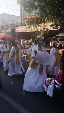 A group of women in traditional white and gold attire are walking in a procession down a street. They are wearing elaborate headdresses and have their faces painted. The street appears to be lined with shops and trees. The sun is shining, casting shadows. Some of the women are looking towards the camera, while others are looking forward. A few onlookers can be seen in the background.
