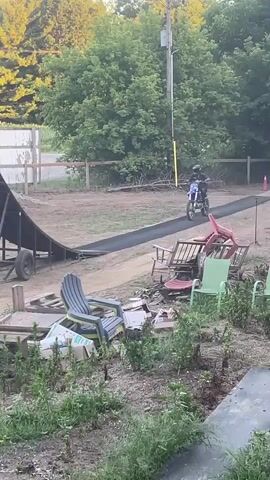 The video shows a person on a dirt bike riding down a ramp, launching into the air, and then landing on an inflatable airbag. The scene is outdoors with trees and a clear sky in the background. There are also other objects visible such as chairs, a trampoline, and some vehicles. The video is shot from multiple angles, with some of the shots being very close to the action. The video appears to be a compilation of a single event, with the action of the dirt bike rider being the main focus.