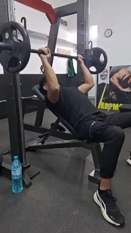 The video shows a man exercising in a gym. He is lying on an incline bench and doing barbell bench presses using a Smith machine. The man is wearing a black t-shirt, black pants, and black and white sneakers. He has short dark hair and appears to be focused on his workout. The gym has black and grey flooring, white walls, and other weightlifting equipment visible in the background. A water bottle sits on the floor next to the bench. A poster with images of bodybuilders is visible in the background.