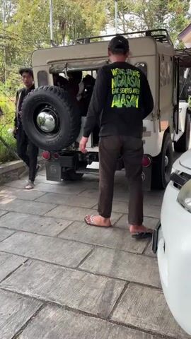 The video opens with a young man in black pants and a tan vest standing by the rear of a vintage off-road vehicle. He has curly dark hair and is looking towards the vehicle. In the background, a second person in a black long-sleeved shirt with yellow writing on the back is standing near the side of the vehicle, facing away from the camera. Inside the open rear of the vehicle, two individuals appear to be lying down. The ground is paved with grey rectangular stones. The scene is outdoors, with trees and foliage visible in the background. As the video progresses, the young man in the vest walks towards the rear of the vehicle, placing a backpack on his shoulders. He then turns and walks towards the side of the vehicle, disappearing from view. The second person remains stationary.