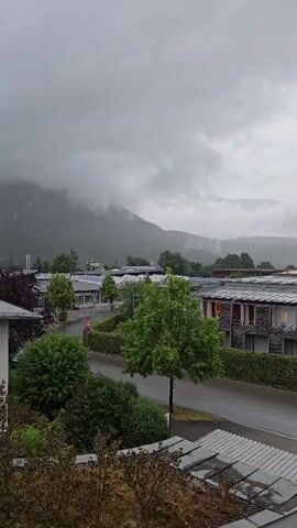 The video opens with a wide shot of a suburban area on a cloudy, possibly rainy day. In the foreground, there are rooftops with some green foliage. As the camera pans slightly, a street with a few houses and buildings comes into view. The houses have pitched roofs and some have solar panels. Trees and hedges are present throughout the scene, adding to the green landscape. In the middle ground, there's a larger building complex with a prominent roof structure, also covered in solar panels. A street sign is visible on the left side of the road. Beyond the buildings, a mountain range dominates the background, shrouded in mist and clouds, giving the scene a moody and atmospheric feel. The overall impression is of a quiet, overcast day in a town situated near a mountainous region.