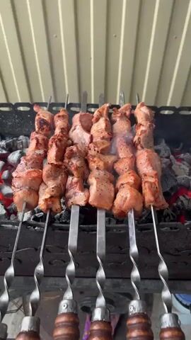 The video shows several skewers of meat being grilled over hot coals. The meat appears to be pork, cut into bite-sized pieces and seasoned with black pepper. The skewers are placed on a metal grill, and smoke is rising from the coals. The background is a light-colored, corrugated metal wall. The lighting suggests it is daytime, likely outdoors. The camera is positioned slightly above the grill, looking down at the cooking meat. There are no people visible in the video.