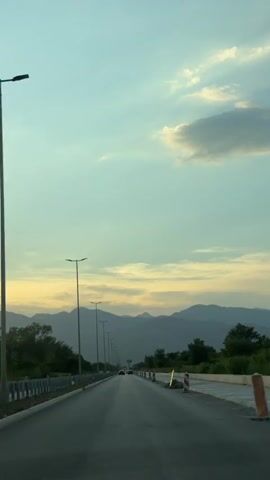 The video shows a view from a vehicle driving on a highway at dusk. The sky is a gradient of pale blue and yellow, with scattered clouds. Mountains are visible in the distance. The highway has multiple lanes and is lined with streetlights and guardrails. Trees and vegetation are on either side of the road. Several cars are also on the highway. The sun is low on the horizon, casting a warm glow. There are some construction barriers on the right side of the road.