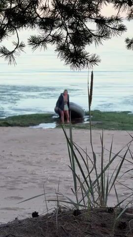 A woman is walking out of the water onto a sandy beach, pulling a gray inflatable boat behind her. She is wearing a dark jacket, a light-colored top, and light-colored shorts. The boat is partially submerged in the shallow water. There is green seaweed or aquatic plants along the edge of the water. In the foreground, there is sand and some tall, dry grass. In the background, the calm water of a lake or ocean stretches to the horizon, with some trees visible at the top edge of the frame. The lighting suggests it is either early morning or late evening.