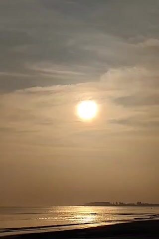 The video shows a sunrise or sunset over the ocean. The sun is visible as a bright disk in the sky, partially obscured by thin clouds. The sky is a mix of light gray and golden hues. Below the sky is the ocean, with gentle waves lapping onto a dark sandy beach. A low-lying landmass with trees is visible in the distance. The scene is still and tranquil.
