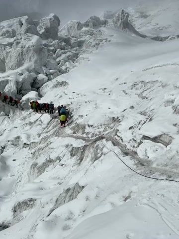 A group of climbers are ascending a steep, snow-covered glacier. They are roped together and using climbing gear, including ice axes and crampons. The terrain is challenging, with large ice formations and crevasses. The sky is overcast, and the overall atmosphere is one of cold and harsh conditions.