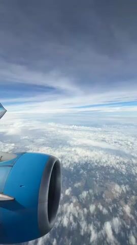 This video shows a view from an airplane window looking out at the sky and the ground below. In the foreground, part of the airplane's engine is visible, with its bright blue casing and dark opening. Above the engine, a portion of the airplane's wing with a light blue tip is seen. The sky is filled with clouds of varying densities, some white and fluffy, others wispy and grey. Sunlight filters through, creating a bright, airy atmosphere. Below the clouds, the ground is visible as a patchwork of greens and browns, suggesting a landscape with fields and possibly some sparse vegetation. The overall impression is one of flight and vastness.