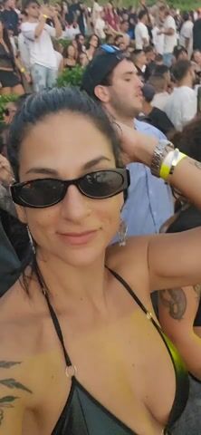 Here's a description of the video:

The video shows a woman taking a selfie in a crowded outdoor setting. She has dark hair pulled back, sunglasses, and is wearing a black bikini top. She smiles at the camera.  Behind her is a dense crowd of people. She adjusts her hair with one hand. The general setting appears to be some kind of outdoor party or festival, with music playing.  Another person enters the frame and smiles at the camera. Other people in the background dance and mingle.