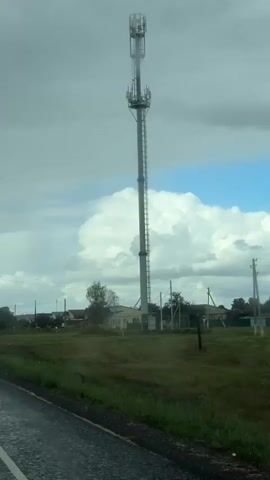 The video shows a tall cell tower with antennas at the top, situated in a rural or suburban area. In front of the tower is a field with some grass and dirt. Beyond the field, there are houses and other buildings, suggesting a small settlement. Several utility poles with wires are also visible. The sky is cloudy with some patches of blue. The camera appears to be mounted on a vehicle, as evidenced by the motion blur and the perspective of the road in the foreground. There are water droplets on what appears to be the windshield, indicating it may be raining or has recently rained. The overall scene depicts a transition from a paved road to a more rural landscape with infrastructure for communication.