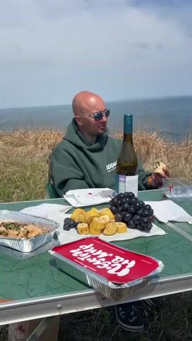Here's a detailed description of the video:

The video shows a picnic scene overlooking a blue ocean on a bright, sunny day. Three people are seated around a green picnic table covered with food and drinks.

The first person, a bald man with sunglasses and a green hoodie, is seated on the left side of the table. He is holding a piece of food in his hand and looking towards the center of the table. A bottle of wine stands near him.

Across from him, a man with a green cap and a light-colored shirt smiles and gestures with his hands while holding a sandwich. He looks as if he's speaking.

On the right side of the table, a person wearing sunglasses and a tan jacket sits. Their back is toward the camera.

The picnic table holds grapes, sliced bananas, containers of prepared food, and paper cups. White napkins are scattered around. A red container with white lettering sits near the edge of the table closest to the camera.

The table is situated on a grassy hillside. The ocean stretches to the horizon, with some islands or landmasses visible in the distance. The sky is blue with a few scattered clouds.