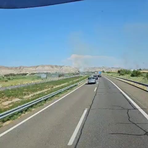 The video shows a view from a vehicle traveling on a highway. The highway is paved with asphalt and has a white line marking the lane. There are guardrails on the side of the road. In the distance, there are hills and vegetation. There are a few cars on the highway. There is also smoke in the distance. The sky is clear and blue. The video is short and does not show any people.
