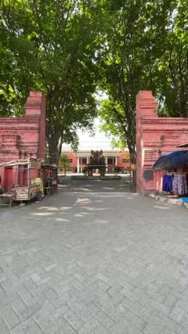 A static shot from a low angle looks through a stone gateway with pink walls and ornate carvings. Two vendors with carts are visible on either side of the gateway. Beyond the gateway, a courtyard is visible with a building and a statue at the far end. Trees with lush green foliage form a canopy over the courtyard. A person rides a motorcycle through the courtyard.