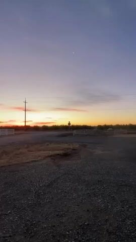 The video shows a wide shot of a rural landscape at dawn or dusk. The sky is a gradient of purple, pink, and orange, with a crescent moon visible high up. A utility pole stands tall in the middle ground, with power lines stretching across the sky. A white fence runs along the left side of the road, and a stop sign is visible on the right. The foreground is a gravel area, and a portion of a vehicle is visible at the bottom left. The overall impression is one of peaceful, natural beauty.