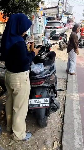 Two young women wearing hijabs stand next to a motorcycle parked on the side of a street. One woman, on the left, is wearing a dark blue hijab and tan pants. She is holding a cup and looking down at her phone. The other woman, slightly to her right, is wearing a dark green shirt and tan pants, and is also looking at her phone. She smiles at the woman on the left. Another woman, wearing a light brown hijab and white pants, stands further down the street to the right. She is looking off to the side. The motorcycle is black and has a license plate that reads A 5353 DD. A white plastic bag is on the seat of the motorcycle. The background shows buildings, other motorcycles, and a car. The ground is dirt and asphalt.