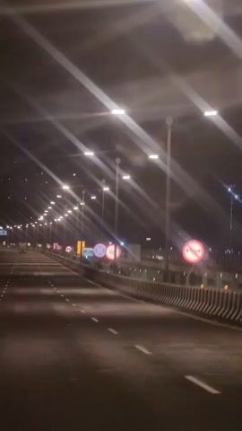 The video shows a nighttime view of a multi-lane highway. Streetlights illuminate the road, casting long streaks of light. Traffic signs are visible along the side of the road, including speed limit signs and other regulatory signs. The road is mostly empty, with only a few distant vehicles visible. The overall atmosphere is dark and somewhat deserted.