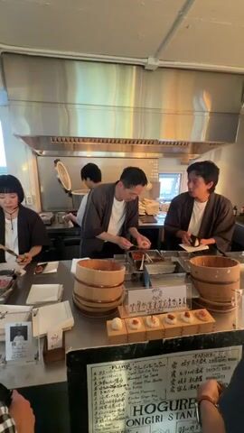 Three individuals are working behind a counter in what appears to be a food preparation area, possibly a small restaurant or food stand. The person in the center is actively preparing food, perhaps dipping it into a liquid from a container. The person on the left is also handling food items. The person on the right is writing or taking notes while holding a food item. The menu is clearly displayed, containing Japanese characters and English descriptions, and the name "HOGURE STAND" is visible. There are wooden buckets, a tray of prepared food items, and other food preparation tools on the counter. The environment is well-lit with a large stainless steel hood overhead. The focus is on the people preparing food.