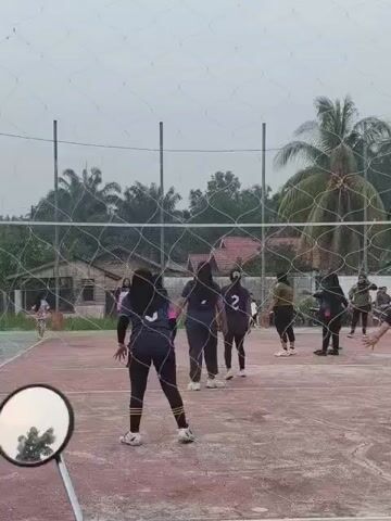 This is a video of a volleyball game being played outdoors on a court. The players are women wearing dark uniforms with numbers on them. One player in the foreground is wearing a dark hijab and dark leggings with yellow stripes. Another player in the background is wearing a dark uniform with the number 2 on it. The net is visible, and the ball is in play. There are palm trees and houses in the background. The sky is overcast. In the lower left corner of the frame, a motorcycle's rearview mirror is visible, reflecting a tree.