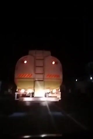 The video shows the rear view of a large tank truck driving on a road at night. The truck is light-colored, possibly white or cream, with prominent red tail lights and amber reflective chevron stripes across its back. A ladder is visible in the center of the truck's rear. The truck's undercarriage is illuminated by its own lights. The scene is dark, indicating it is nighttime, and the road ahead is visible as a dark surface. The video is likely filmed from a vehicle following behind the truck in Kuta Pungwa, Indonesia. No specific activity beyond driving is discernible.