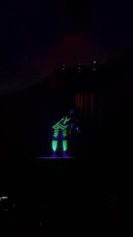 A lone performer stands on a lit platform in a dark venue, wearing a costume with neon green geometric lines. The performer is illuminated by a bright green laser beam shining from above and a sweeping purple laser beam crossing the stage from right to left. The background is dark with faint red curtains visible. The performer's posture and the lighting suggest a performance or show.