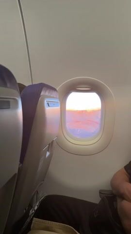The video shows the view from an airplane window. Inside, you can see a purple airplane seat in the foreground, and the rounded frame of the window beside it. Outside, the sun is on the horizon, with golden light shining through a layer of clouds. The sky is a mix of blue and pink, and there appears to be water or more clouds below. The window is slightly smudged. Part of an arm is visible in the bottom right corner of the frame.