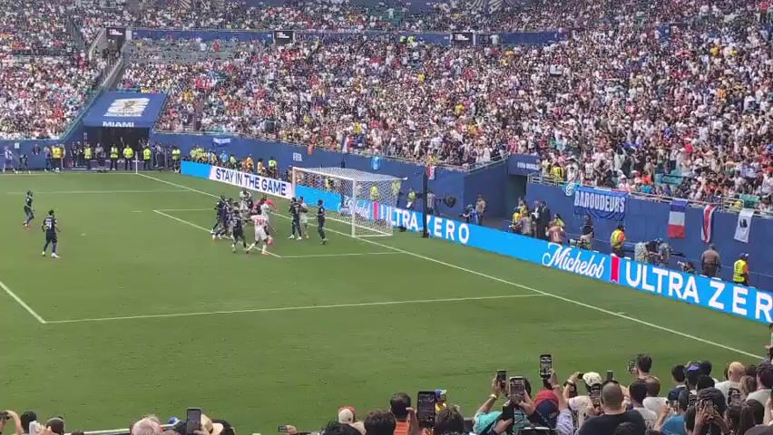 A soccer match is underway in a large stadium filled with spectators. The field is green with white markings. Two teams are on the field, one wearing white uniforms and the other dark blue. The white-uniformed team is clustered around the goal, possibly during a penalty kick or corner kick. The dark blue-uniformed team is defending. Spectators are visible in the stands, many holding up cell phones to record the game. Advertising banners are placed along the edge of the field. The sky appears bright, suggesting it is daytime.