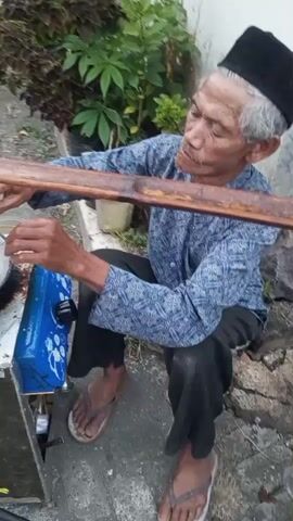 An elderly man wearing a blue patterned shirt, dark pants, and a black cap is sitting on the ground next to a portable stove setup. He is holding a wooden stick, using it to stir or move food in a frying pan. The frying pan is placed on top of the stove, which is part of a metal cart. The cart has a blue decorative panel with white floral patterns. The man is outdoors, with plants and a stone wall visible in the background. His feet are clad in flip-flops. The camera is positioned at a medium distance, capturing the action and the immediate surroundings.