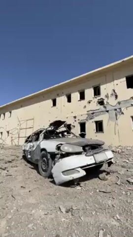 The video shows a badly damaged car in front of a building. The car is white and appears to have been burned and severely damaged, with its roof partially collapsed. The building behind the car is beige and has numerous bullet holes and other damage. The area around the car is covered in debris and rubble. The sky is clear and blue. There are indistinct spoken voices in the background.