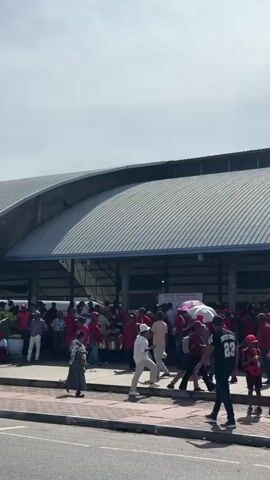 The video shows a crowd of people gathered on a brick-paved area in front of a large building with a curved metal roof. The majority of people are wearing red shirts or hats, and some are carrying small red flags. A few people hold umbrellas, likely for shade. The crowd appears to be loosely organized, with individuals standing and walking around. To the left of the frame, a sign indicates "Food City DELKANDA 02" and a distance of 500 meters. The sky is partially cloudy. A black water tank on a green metal stand is visible within the crowd. Some individuals are interacting near the water tank. Cars can be heard driving nearby.